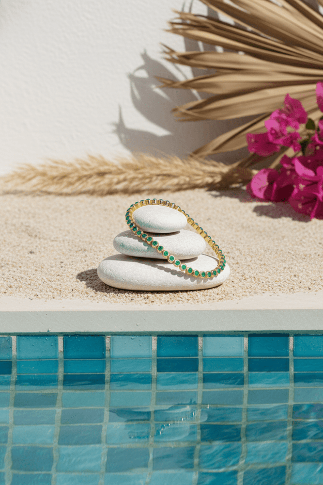 
                  
                    Bracelet with green stones on a stack of stones by a poolside with flowers and palm leaves.
                  
                