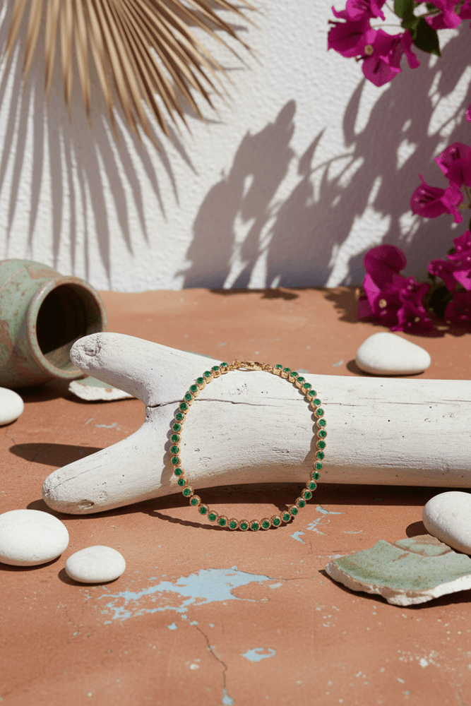 
                  
                    Green tennis bracelet on a wooden branch with pink flowers and pebbles in the background
                  
                