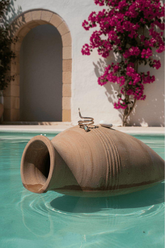 
                  
                    Decorative vase with a spiral design floating in a pool with a white building and pink flowers in the background.
                  
                